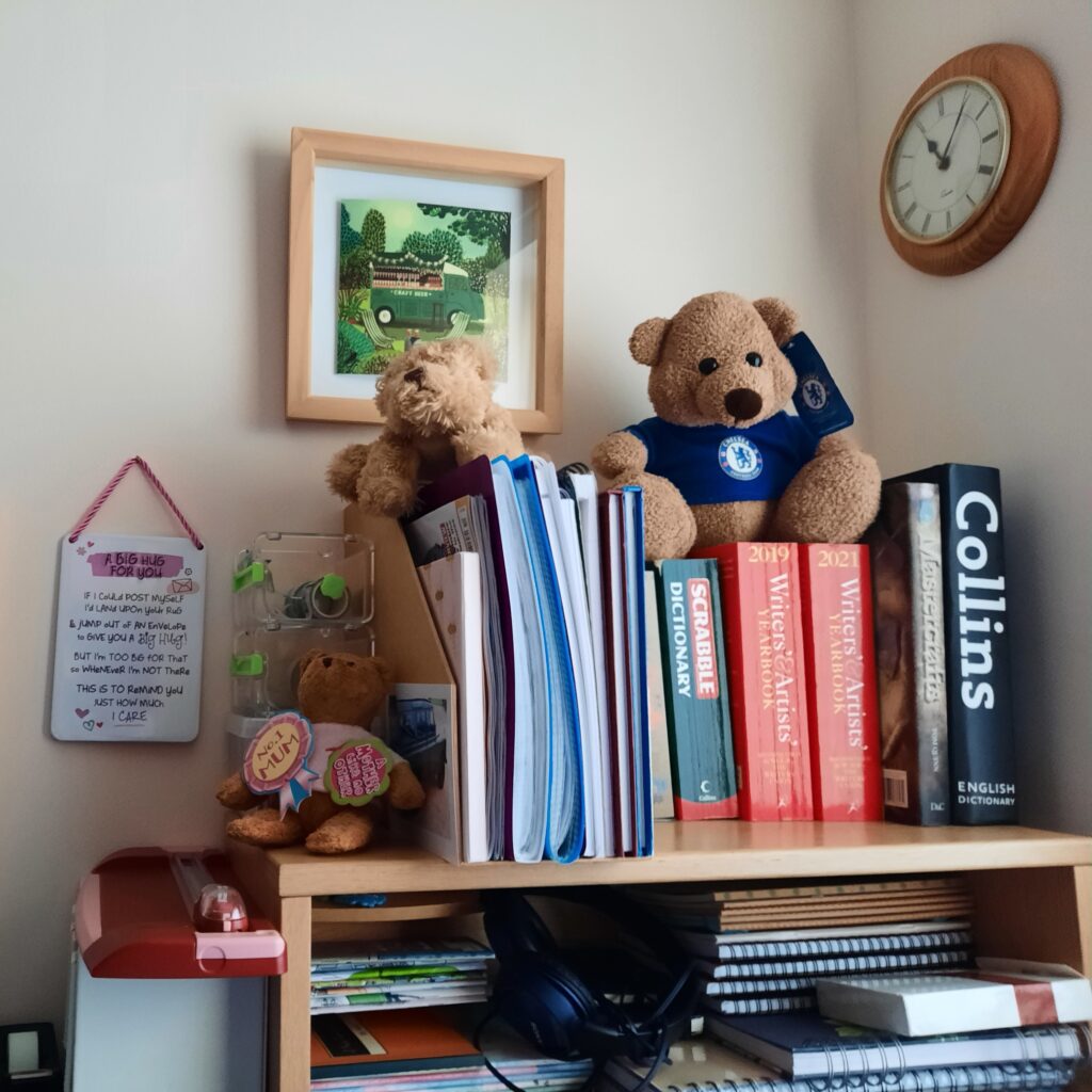 This photograph shows a shelf that sits on top of the author's desk. Items on the shelf include a Collins Dictionary, 2 copies of the Writers & Artists Yearbook, one is 2019, the other is 2021, both are red in colour, and a SCRABBLE Dictionary that is now too old to rely upon, so we bought the Collins Dictionary. 

Various notebooks are held in a manila card magazine file storage case.

There are 3 stuffed toys including a small teddy bear who proudly wears a No 1 Mum badge. The Chelsea Football Club mascot and a fluffy dog that cleans my laptop screen are also precious creatures on my desk.

Plaques and pictures adorn the plain beige wall. There's an analogue clock that reads 5 minutes past 11, or 1105 in digital format.
