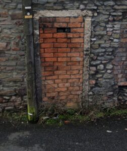 Images show two views of the end wall of a building where a doorway has been blocked out. One brick is missing, rather like a posting or spy hole has been left.